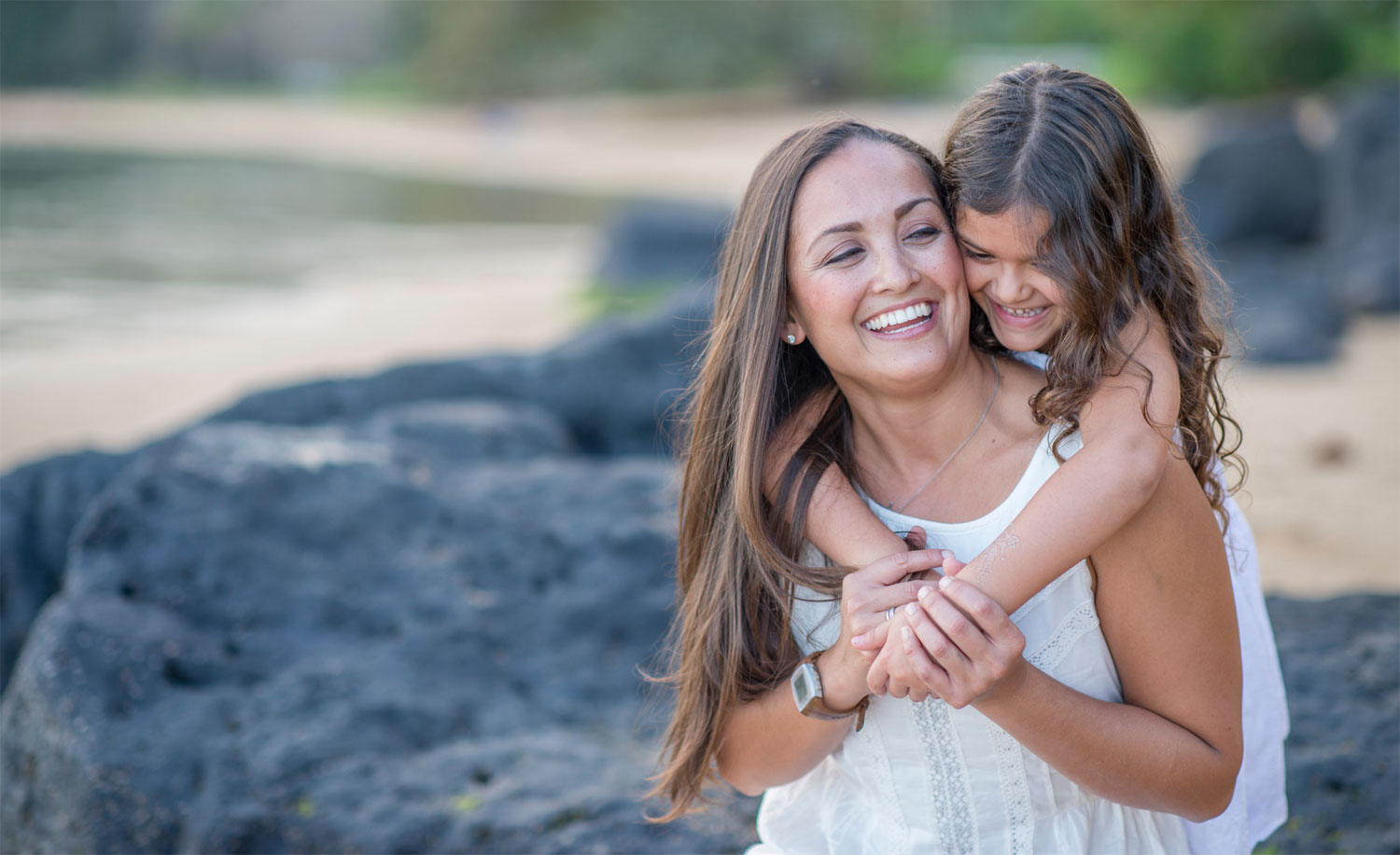 A happy mom and girl in white sleeveless dress cuddling each other in front of the beach background