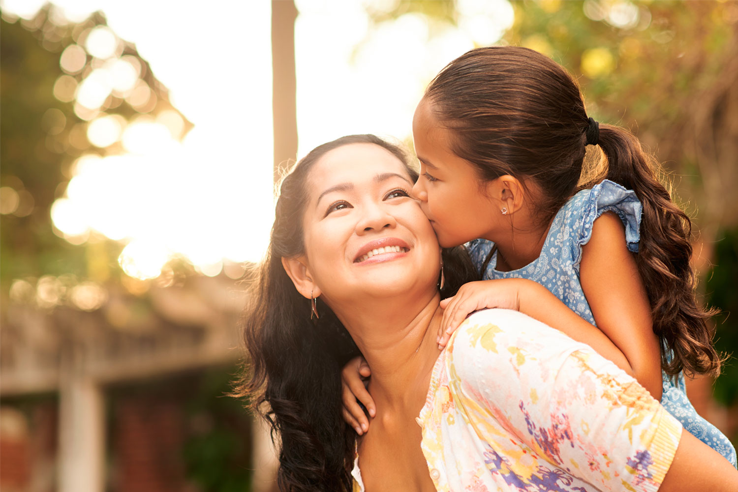 A happy mom in pattern cardigan carrying a girl on her back in a bright sunny day background